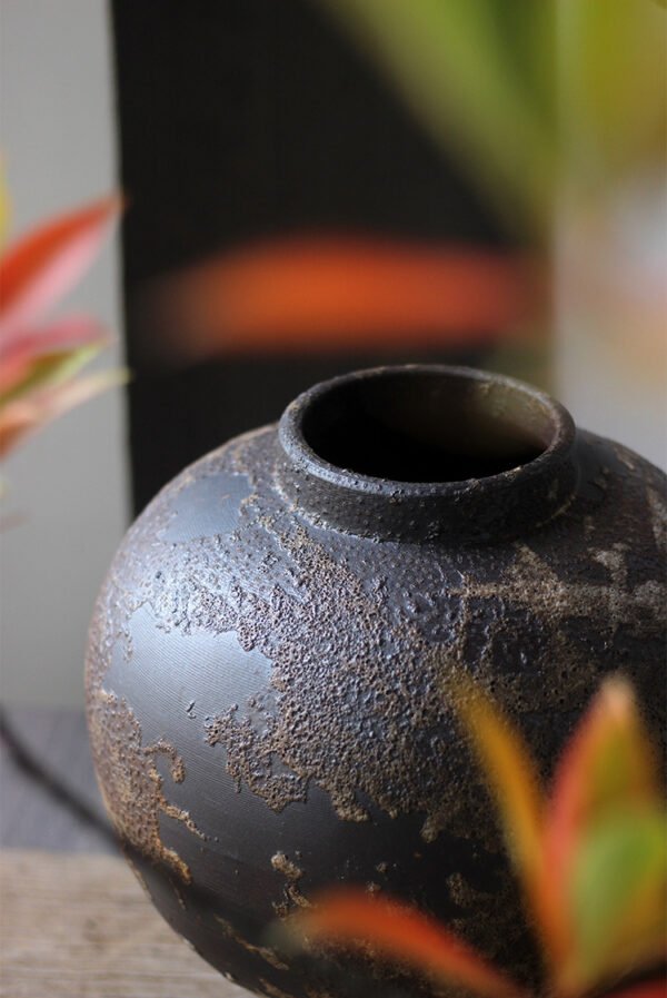 Textured dark ceramic vase with blurred plants